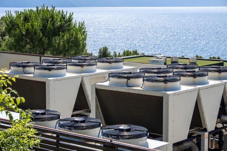 Rows of large commercial HVAC units on a rooftop, strategically positioned for optimal air circulation and cooling efficiency. Overlooking a calm sea, merging industrial technology with natural beauty. Ideal for themes of sustainability and energy management in technology.