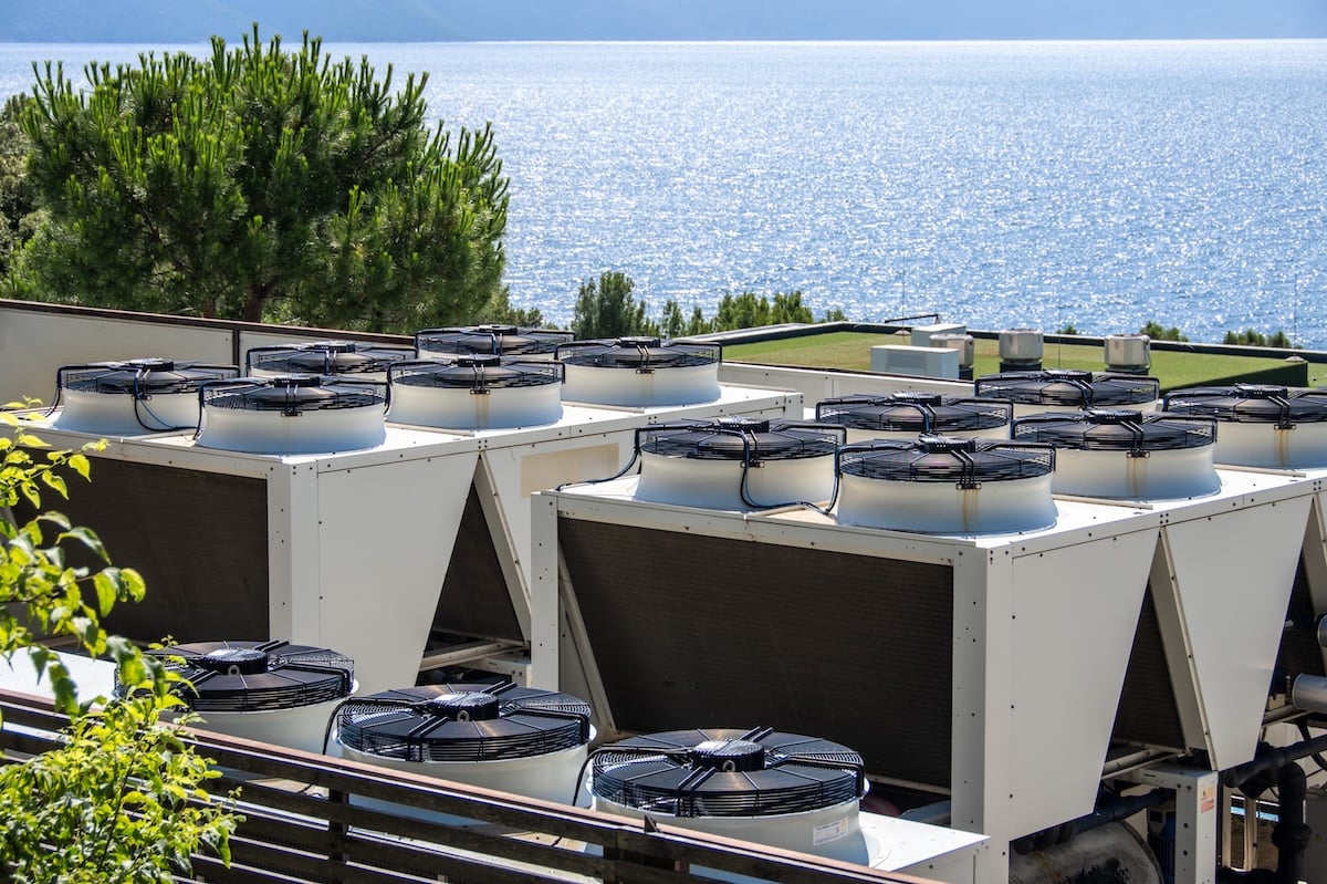 Rows of large commercial HVAC units on a rooftop, strategically positioned for optimal air circulation and cooling efficiency. Overlooking a calm sea, merging industrial technology with natural beauty. Ideal for themes of sustainability and energy management in technology.