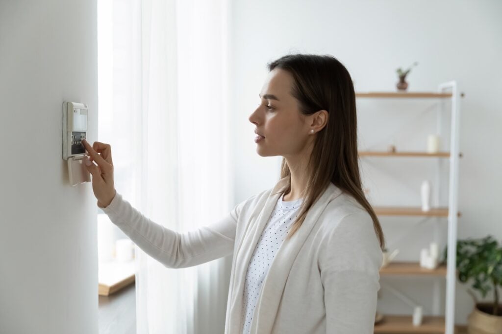 how to save money on heating bill Side view pleasant young woman using smart home system or activating modern alarm system before leaving apartment. Happy lady turning off easy security technology, when returning house or flat.