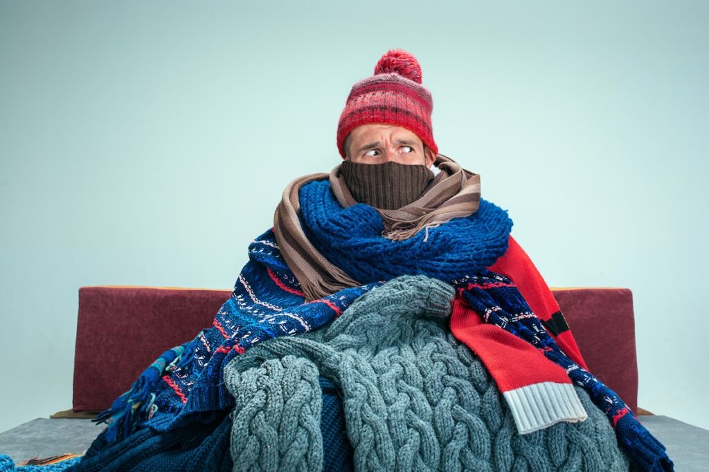 how to keep house warm in winter Bearded sick man with flue sitting on sofa at home or studio covered with knitted warm clothes. Illness, influenza, pain concept. Relaxation at Home. Healthcare Concepts.