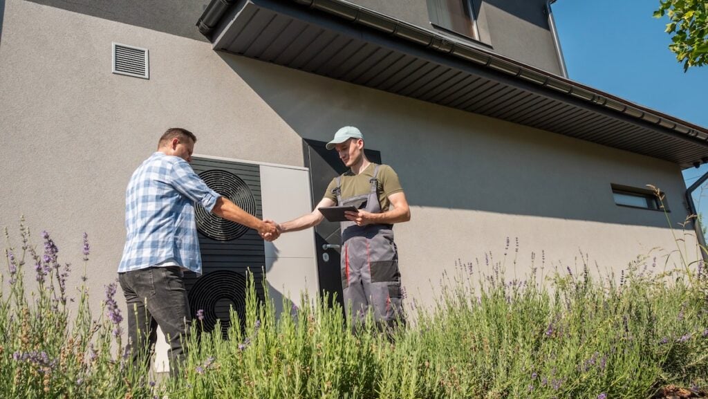A technician and a homeowner discussing heat pump maintenance outside a house